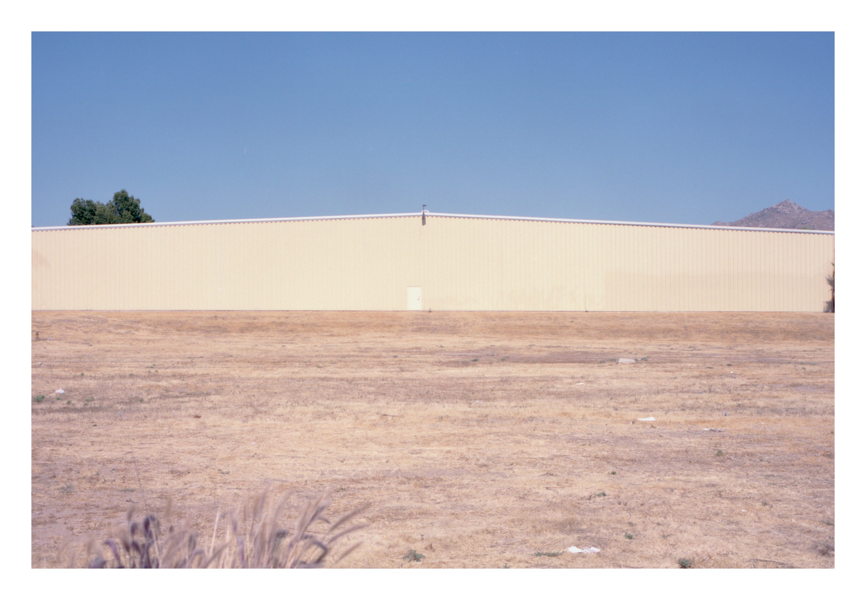 A long pale-cream warehouse wall stretching across the frame against a deep blue sky, with a single small door barely visible at the wall's base, dry desert grass in the foreground, and distant low mountains on the right.