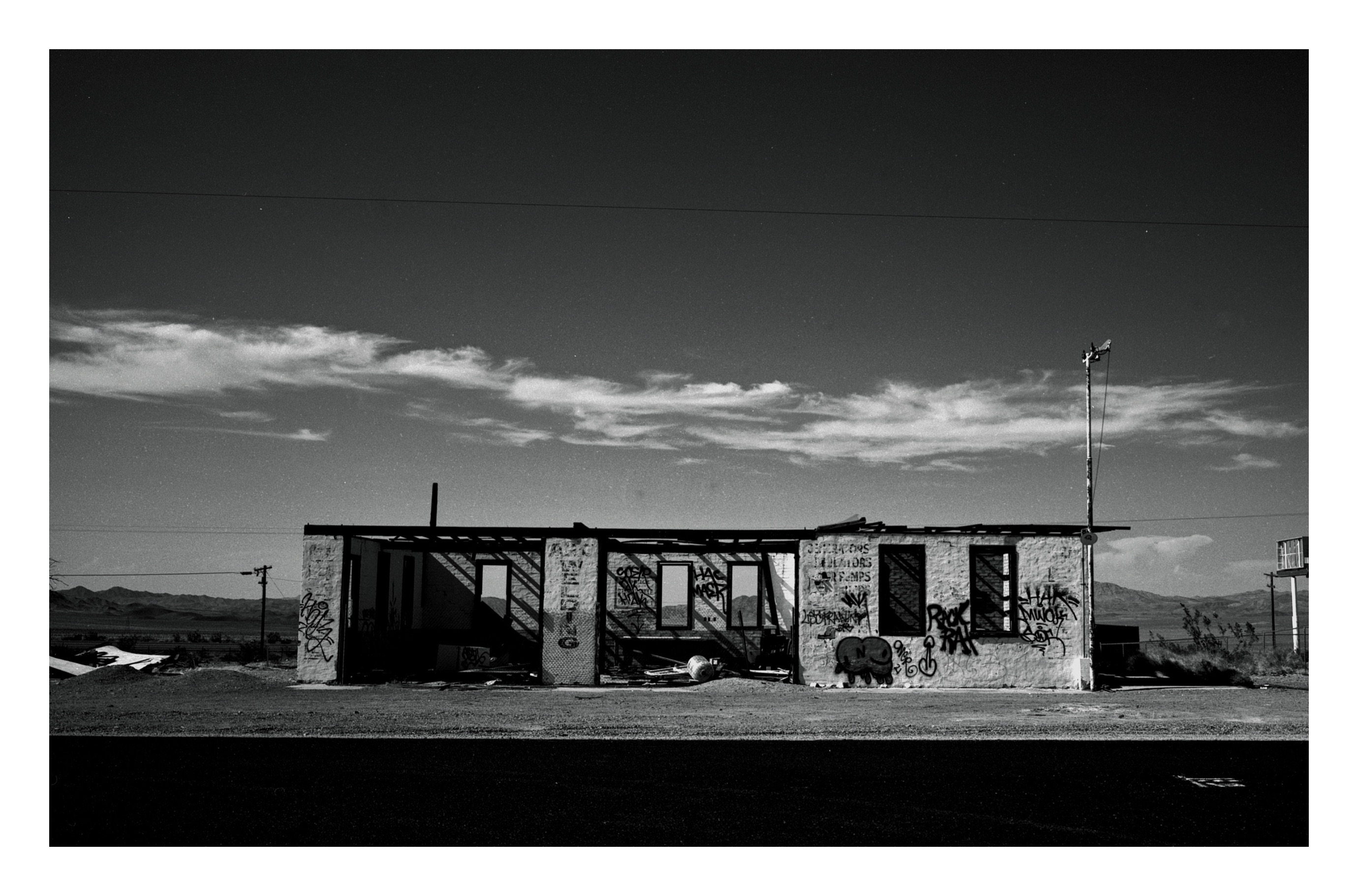 A roofless graffiti-covered cinder-block building in the open desert, low mountains and power lines in the distance, asphalt road in the foreground.