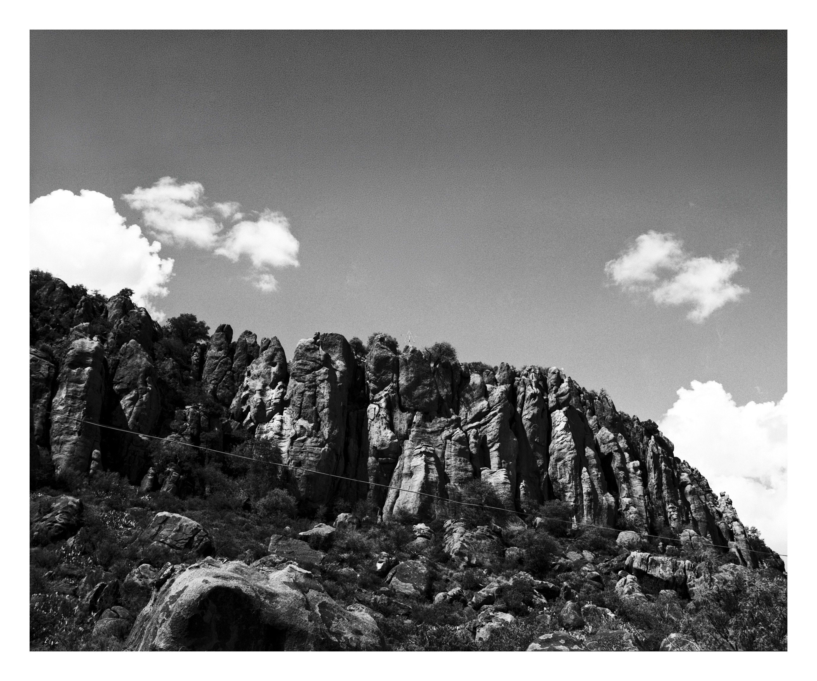 A long ridge of vertically folded rock columns with desert scrub at the base, rising under a sky of cumulus clouds.