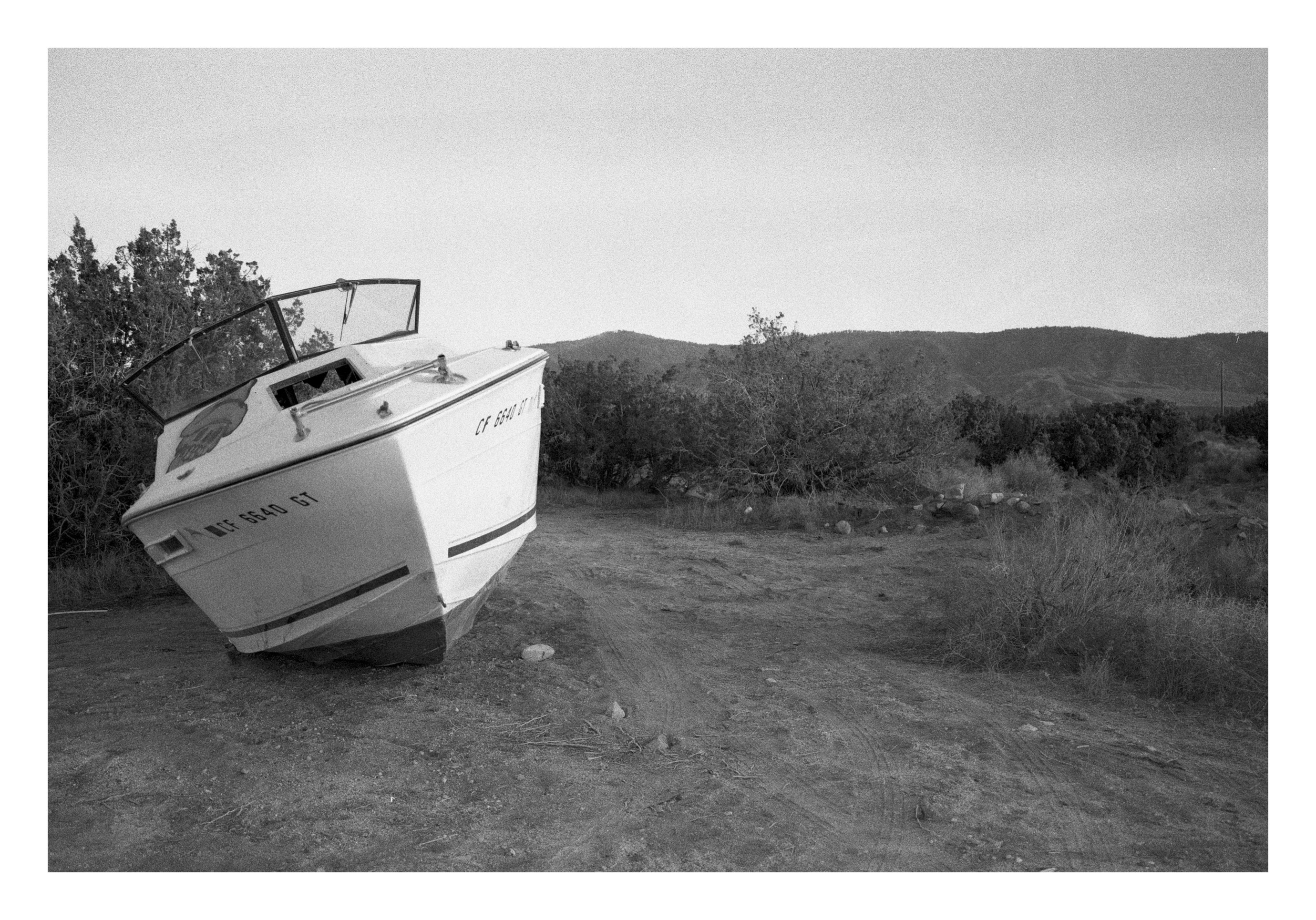 A small motorboat tilted on bare desert dirt with a partial California ID number on the hull, scrub and low mountains behind.