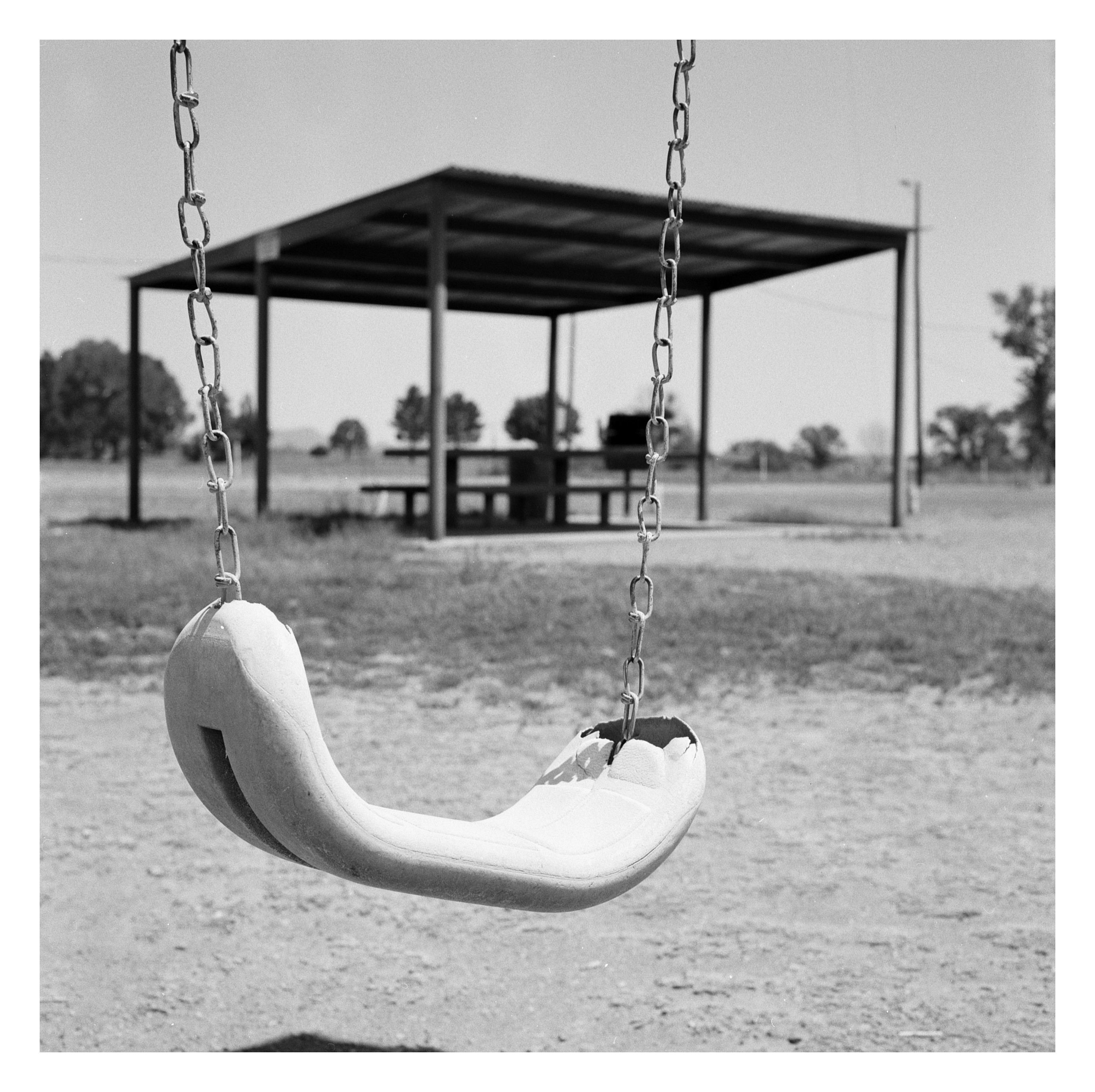 A black-and-white photograph of an empty playground swing hanging on chains in the foreground, with a covered pavilion and distant trees in the background.