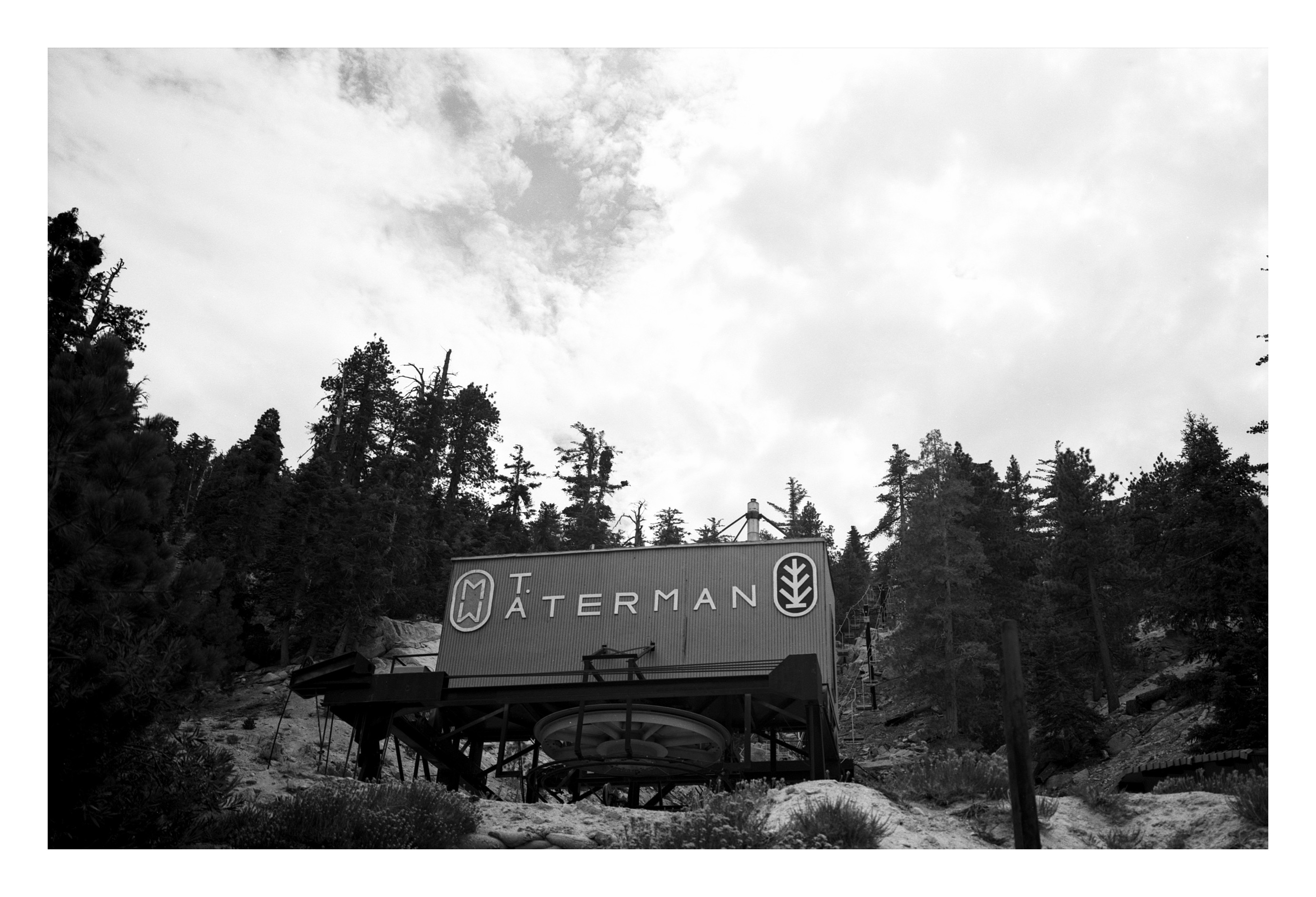 A weathered ski-lift loading station with the MT WATERMAN logo visible on its side, surrounded by tall pines under a heavy cloud-filled sky.