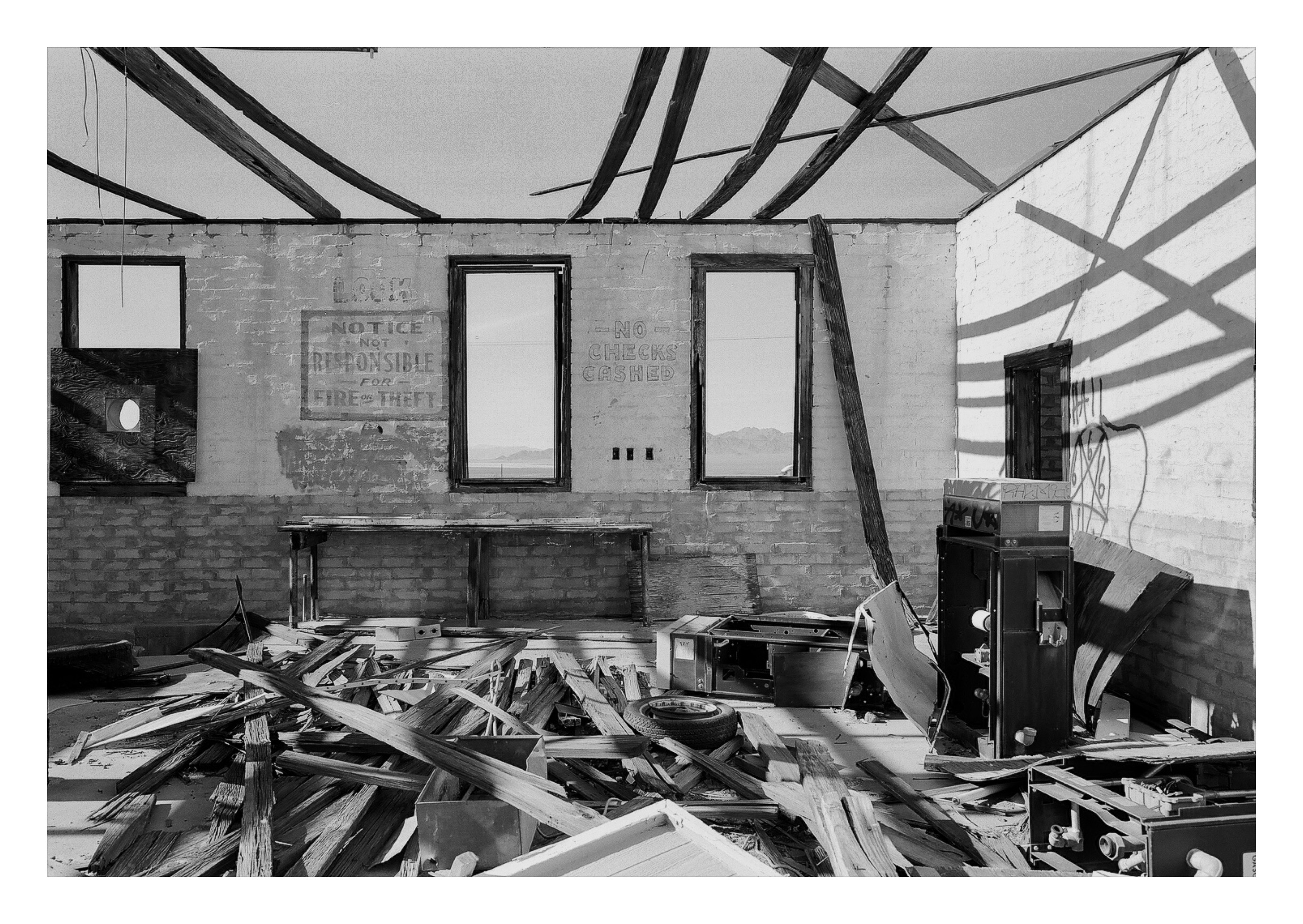 The interior of an abandoned brick building with a collapsed roof, faded signs reading NOTICE NOT RESPONSIBLE FOR FIRE OR THEFT and NO CHECKS CASHED, a vending machine and pile of debris on the floor.