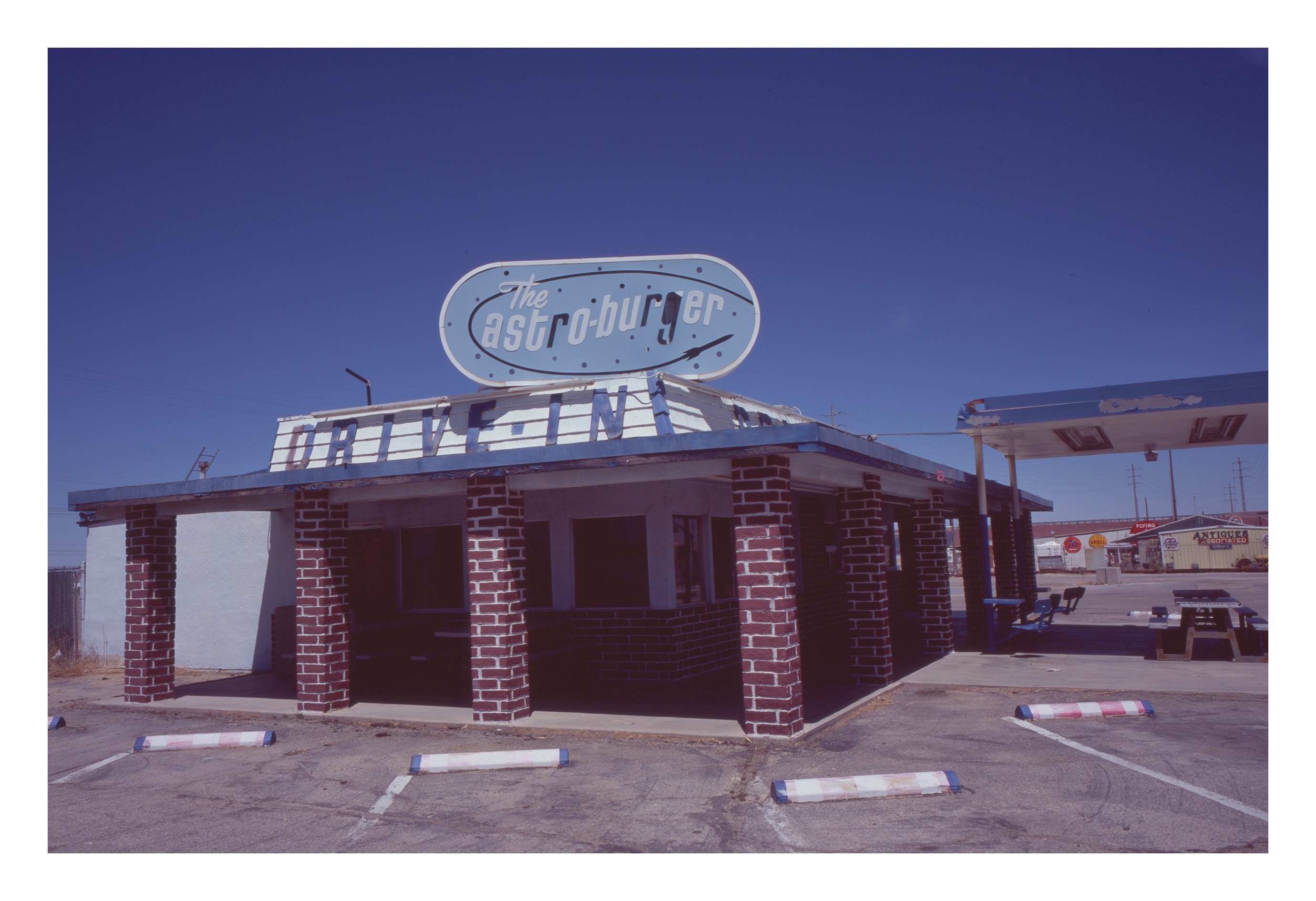 A closed Astro-burger drive-in restaurant on Route 66 with red brick columns, classic Americana signage on a saturated deep blue sky, parking-stop curbs in the empty lot.