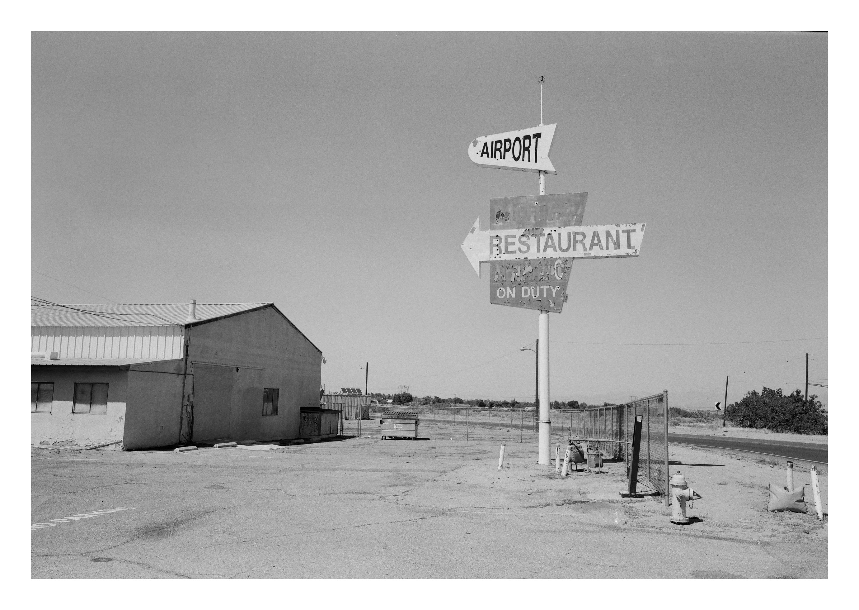 A leaning two-arrow road sign reading AIRPORT and RESTAURANT NO ONE ON DUTY against an empty parking lot, low building, and chain-link fencing.