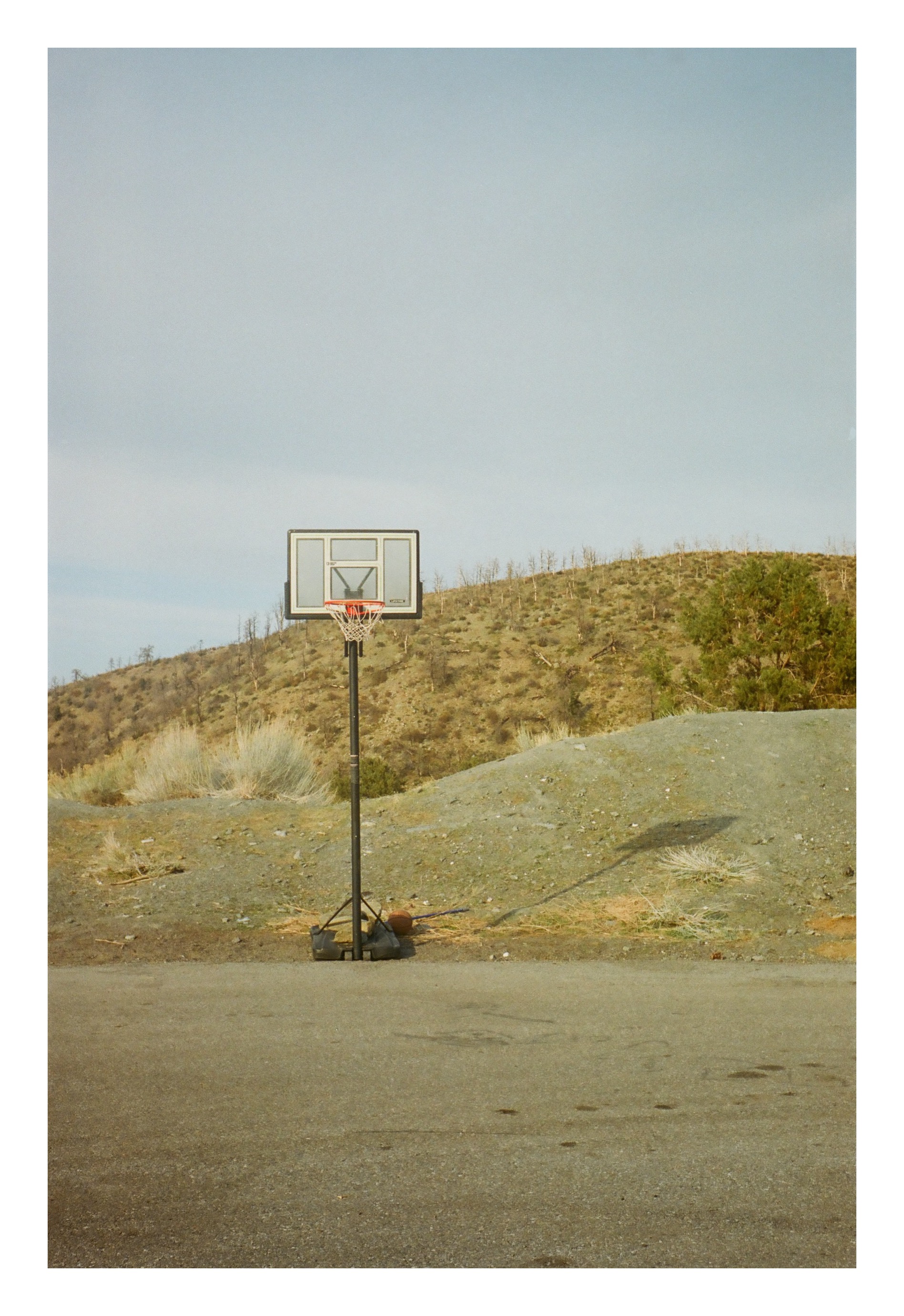 A basketball hoop standing alone on an asphalt court at the foot of a scrub-covered hillside.